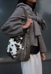Persoon in een bruine trui en beige broek, met een grote schoudertas van leer met een bruin en wit koeienhuidpatroon.