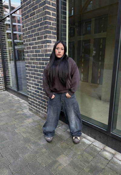 Sudadera gris oscuro de gran tamaño combinada con pantalones vaqueros azules desgastados y holgados. Zapatillas informales completan el look. Paseo pavimentado y pared de ladrillo de fondo.