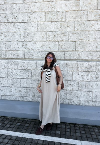 Mujer con vestido maxi beige y suéter de rayas colocado sobre los hombros, de pie frente a una pared de piedra clara en una acera pavimentada.