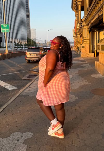 Vestido rosa de cuadros vichy con bolsillos, combinado con zapatillas Nike blancas y naranjas y gafas de sol rosas. Vista lateral, fondo urbano.