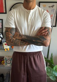 White short-sleeve henley shirt with three buttons, paired with brown athletic trousers featuring side stripes and an elastic waistband.
