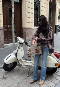 Scooter vintage bianco accanto a una persona che indossa una maglietta a righe marroni a maniche lunghe e jeans blu, mentre tiene una borsa beige con accessori.