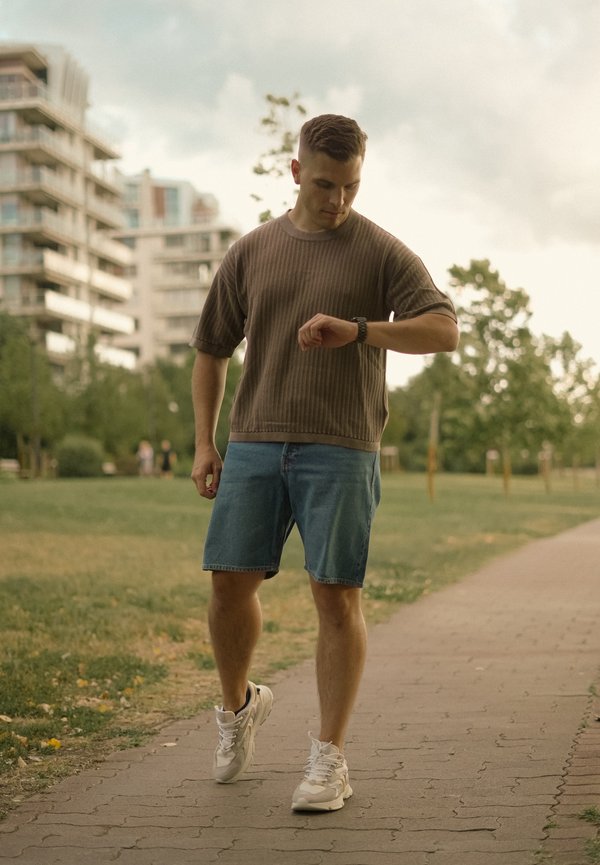 Jeune homme en chemise marron et short en jean marchant sur un chemin de parc, regardant sa montre connectée, avec des bâtiments et des arbres en arrière-plan.