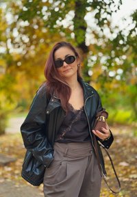 Melna ādas bombers jaka virs melnas mežģīņu topa, bēšas augstās jostas bikses, brūna somiņa ar zelta akcentiem un saulesbrilles.