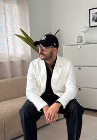 Persona sentada en un sofá beige, vistiendo un blazer blanco sobre una camisa negra y pantalones negros; gafas de montura y una gorra negra; entorno interior con plantas.