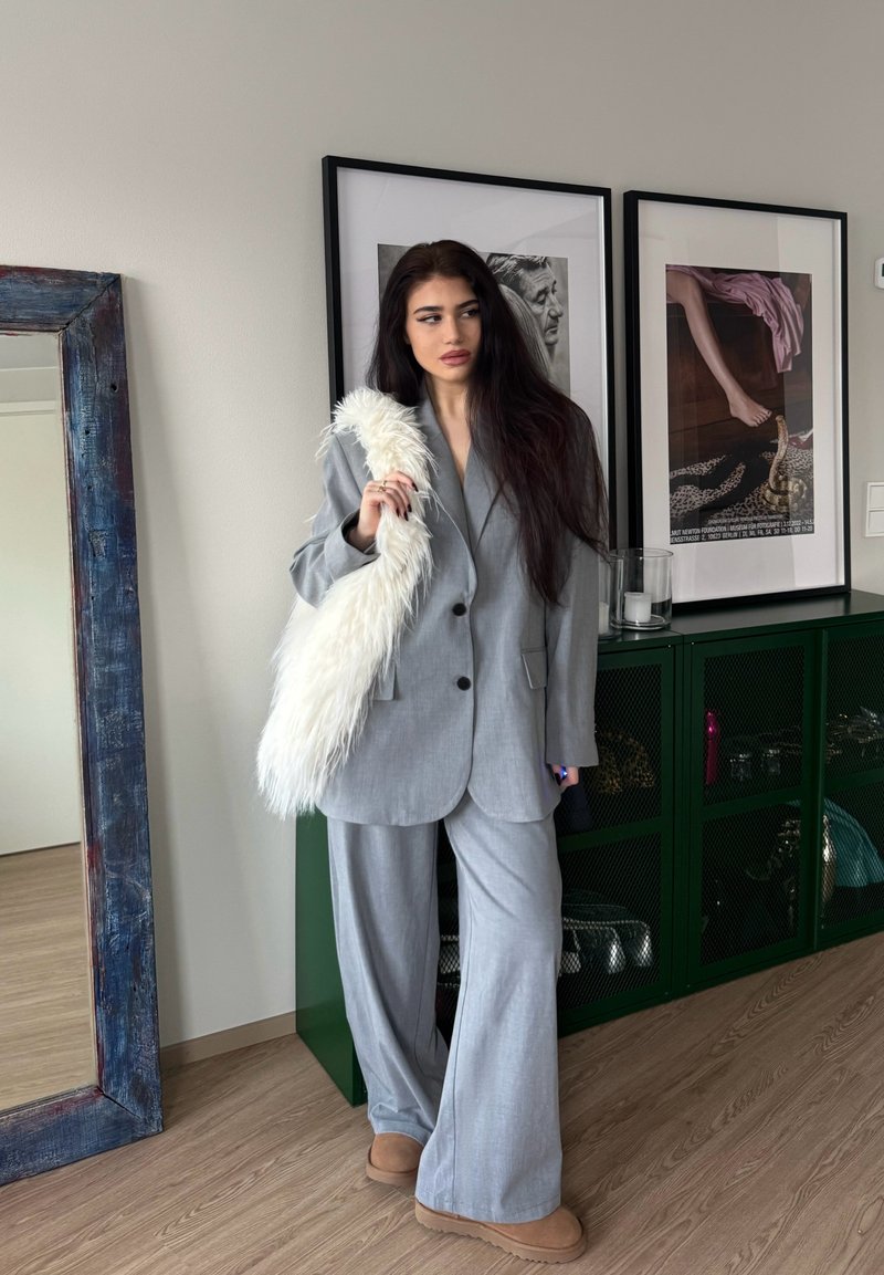 Jonge vrouw met lang donker haar in een grijs oversized pak en beige schoenen, met een witte, pluizige tas in haar hand, staand in een kamer met ingelijste kunst en een groene kast.