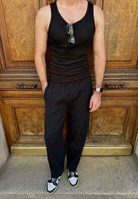 Μαύρο κοντομάνικο μπλουζάκι, μαύρο παντελόνι με ρίγες, και μαύρα αθλητικά με λευκές λεπτομέρειες. Γυαλιά ηλίου που ξεκουράζονται στο λαιμό. Ξύλινο φόντο.