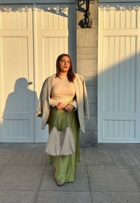 Beige blazer over en creme top og lang grøn nederdel, holder en lys grå håndtaske. Sollyset skaber en blød skygge på gulvet.