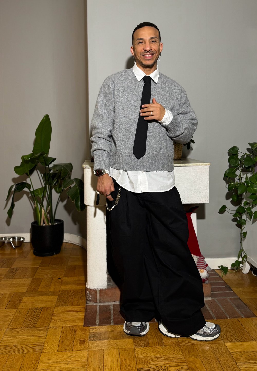 Grauer gestrickter Pullover über einem weißen Hemd, schwarze weit geschnittene Hosen und graue Turnschuhe. Enthält eine Uhr mit Kette und Zimmerpflanzen.