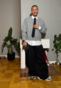 Grauer gestrickter Pullover über einem weißen Hemd, schwarze weit geschnittene Hosen und graue Turnschuhe. Enthält eine Uhr mit Kette und Zimmerpflanzen.