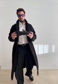 Άνδρας με μαύρο παλτό και γυαλιά ηλίου κρατάει ένα μαύρο παπούτσι μοκασίνι μπροστά σε έναν λευκό τοίχο με ξύλινο πάτωμα.