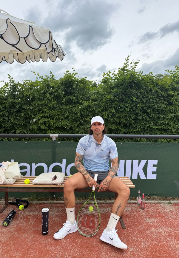 Uomo tatuato in abbigliamento da tennis seduto su una panchina con racchetta e pallina in mano, con attrezzatura da tennis e bevande vicine, cielo nuvoloso sopra.