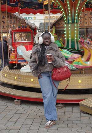 Chaqueta de piel sintética marrón, vaqueros de mezclilla azul de pierna ancha, bolso acolchado rojo, orejeras blancas, sosteniendo una taza de café, de pie junto a un carrusel colorido.