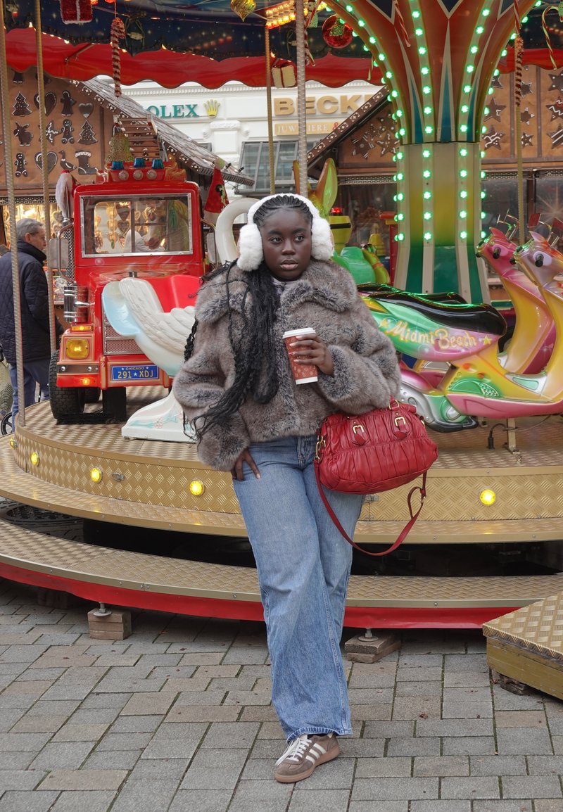 Veste en fausse fourrure marron, jean denim bleu à jambes larges, sac à main rouge matelassé, cache-oreilles blancs, tenant une tasse de café, debout près d'un carrousel coloré.