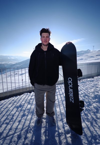 Persona con ropa de invierno sosteniendo una tabla de snowboard negra con la etiqueta "Zalando" de pie sobre la nieve con montañas y una barandilla al fondo.