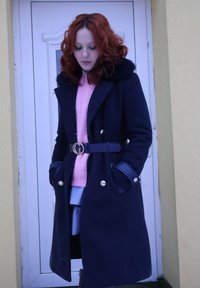 Femme aux cheveux roux bouclés portant un manteau bleu marine avec ceinture sur un pull rose, debout contre une porte blanche et un mur beige.