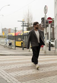 Muž kráčí po dlážděné ulici, má na sobě velký šedý blejzr, bíl�é tričko, černé kalhoty a tenisky, na pozadí je žlutý autobus.