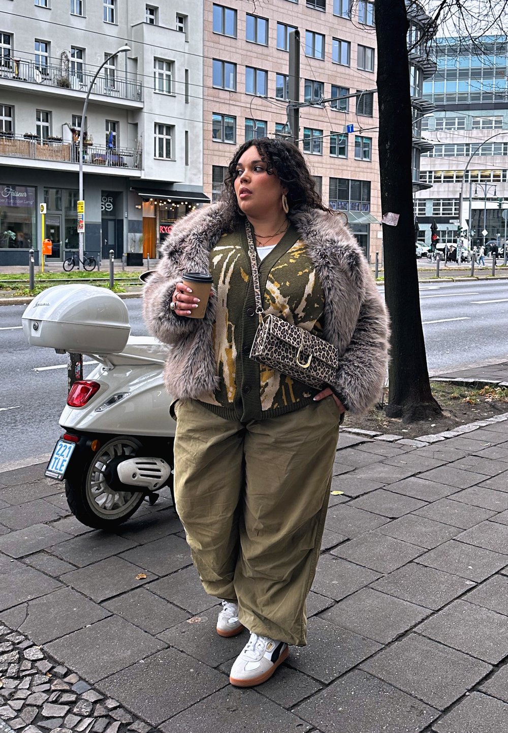 Per didelė pilka dirbtinės kailio striukė, raštuotas alyvuogių žalios spalvos kardiganas, laisvi chaki spalvos kelnės, baltos sportbačiai ir leopardų rašto rankinė. Kavos puodelis rankoje.