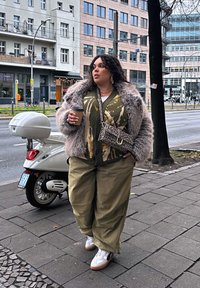 Per didelė pilka dirbtinės kailio striukė, raštuotas alyvuogių žalios spalvos kardiganas, laisvi chaki spalvos kelnės, baltos sportbačiai ir leopardų rašto rankinė. Kavos puodelis rankoje.
