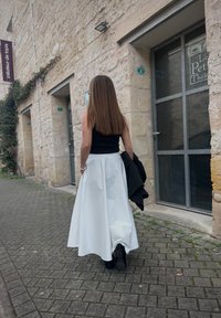 Long white skirt with pockets, black top, and a dark coat draped over the arm. Smooth fabric, flowing design, standing on a cobblestone street.