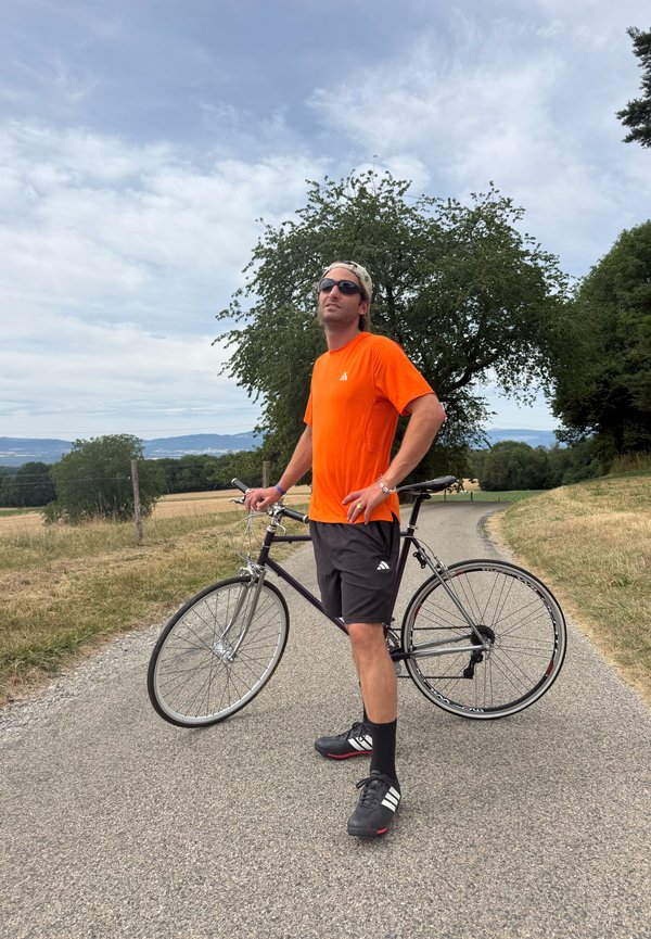Mann in orangefarbenem Hemd und schwarzen Shorts steht mit einem Fahrrad auf einer ländlichen asphaltierten Straße unter einem bewölkten Himmel mit Bäumen und Feldern in der Umgebung.
