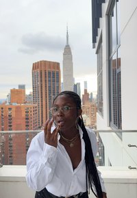 Witte blouse met knopen, gouden kettingen, ronde bril, oorringen in de vorm van hoepels. Stedelijke skyline met het Empire State Building op de achtergrond.