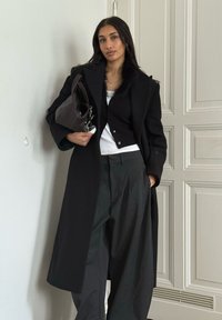 Long black coat with fur accents, paired with a fitted black cardigan, white top, and loose black trousers. Holding a dark handbag.