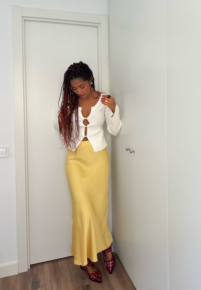 Top de manga larga de canalé blanco con detalle de ojal, combinado con una falda maxi de satén amarilla y zapatos de tacón rojos con tachuelas.