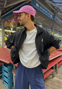 Svart zip-up jacka, vit t-shirt och marinblå shorts. Rosa keps på huvudet. Tillbehör inkluderar silver kedja och örhängen. Bakgrund med marknadsstånd.