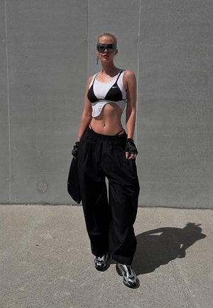 Sujeción deportiva blanca y negra con diseño recortado, pantalones joggers negros holgados, guantes y zapatillas brillantes. El fondo es una pared gris lisa.