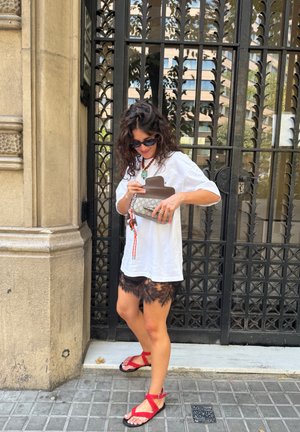 Camiseta blanca de gran tamaño, pantalones cortos de encaje negros, sandalias rojas con tiras y un bolso con estampado y un acento de cadena. Las gafas de sol negras completan el look.