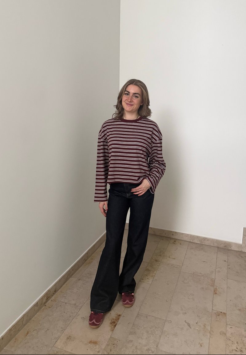 Junge Frau mit schulterlangem Haar, die ein gestreiftes Langarmshirt, schwarze weite Hosen und burgunderrote Sneakers trägt und sich drinnen befindet.