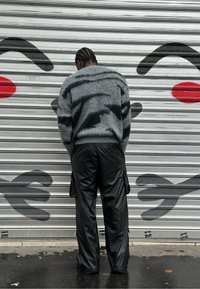 Suéter de punto gris con patrones abstractos negros, combinado con pantalones cargo sueltos de cuero negro. El fondo presenta un rostro pintado sobre una superficie metálica ondulada.