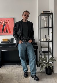 Person wearing a black blazer over a dark jumper and light blue jeans, paired with black shoes. Background features a shelf and artwork.