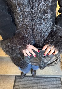 Manteau noir en fourrure avec manches texturées, sac à main noir tenu des deux mains, jeans foncés et bottes noires à bouts pointus. Ongles rouges visibles.