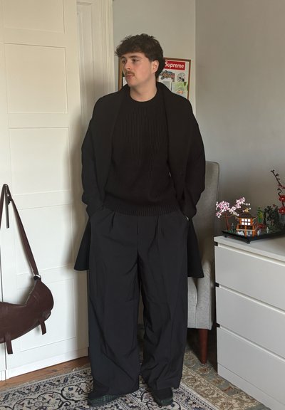 Joven con suéter negro, pantalones anchos negros y abrigo, de pie en el interior cerca de una cómoda blanca con un pequeño modelo de estilo japonés y un tablero de anuncios.