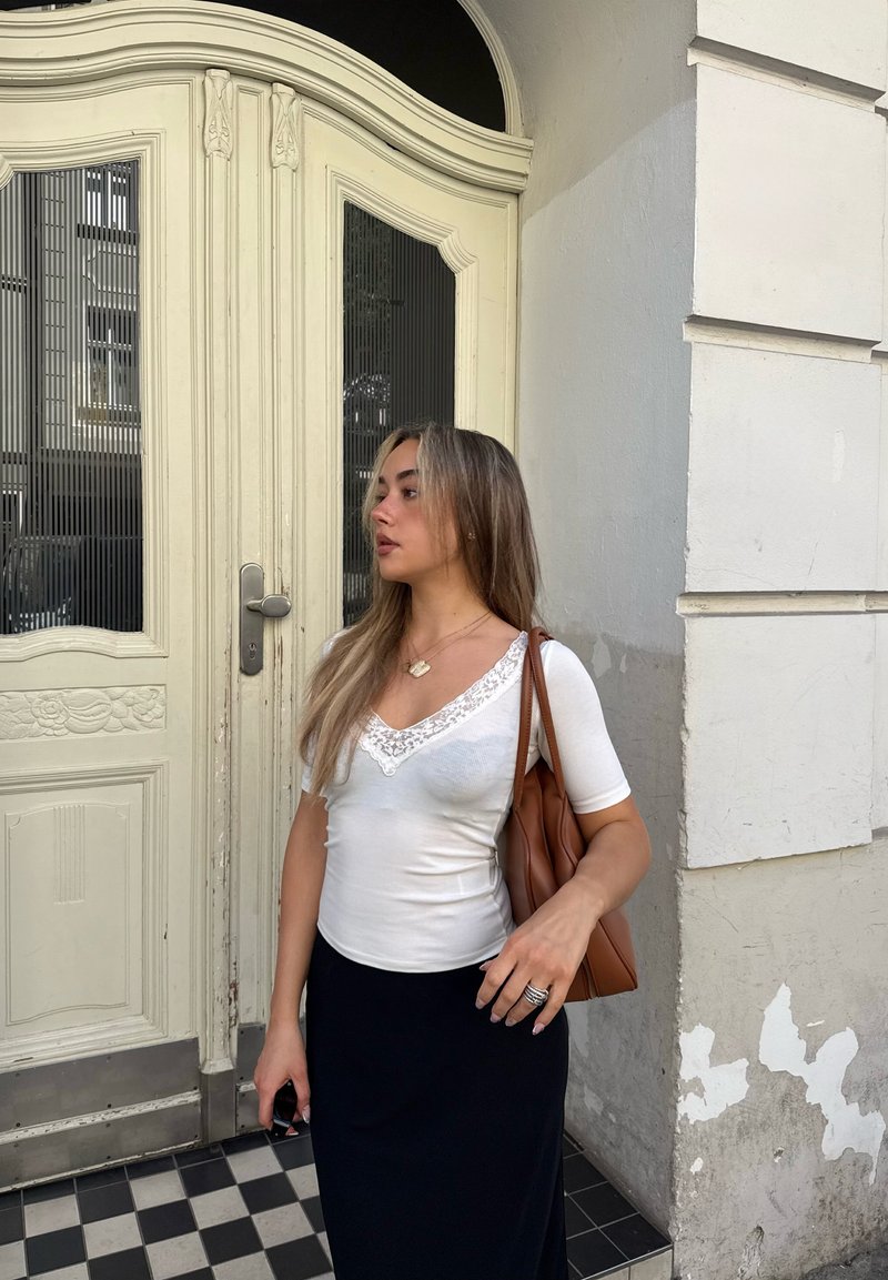 Jeune femme aux longs cheveux portant un haut blanc et une jupe noire, se tient près d'une porte crème ornée de panneaux de verre décoratifs.