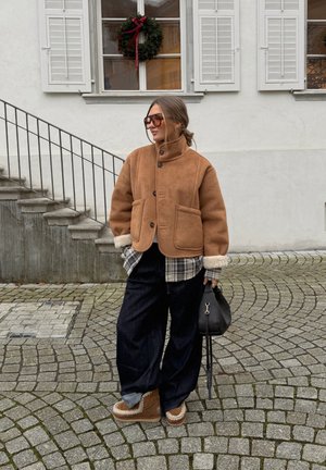 Chaqueta de ante marrón con cuello alto y dos bolsillos delanteros, combinada con una camisa de cuadros. Pantalones anchos de pierna oscura y botas con detalles en crema. Bolso negro.
