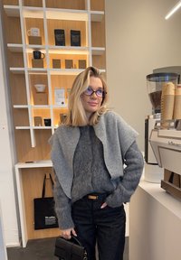 Femme blonde avec des lunettes, vêtue d'un pull gris en tricot et d'un pantalon noir, debout à l'intérieur d'un café près d'une machine à espresso et d'étagères en bois avec des articles de café.