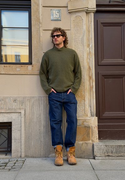 Hombre con cabello rizado y gafas de sol, que lleva un suéter verde, vaqueros azules y botas marrones, está apoyado contra una pared beige junto a una ventana y una puerta.