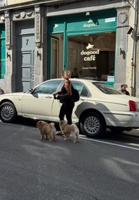 Žena v čiernom prechádza s dvoma malými nadýchanými psami pri zaparkovanom smotanovom aute pred kaviarňou dogood s zeleným výkladom.