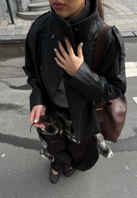 Sort læderjakke med høj krave, grå top, ternet skjorte, brun taske og mørke sko. Holder mørke solbriller, hænder prydet med ringe.