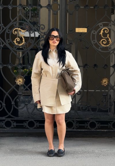 Mujer con cabello largo y negro y grandes gafas de sol lleva un vestido camisa beige de gran tamaño, sosteniendo un bolso marrón, de pie frente a una ornamentada puerta de hierro negra.