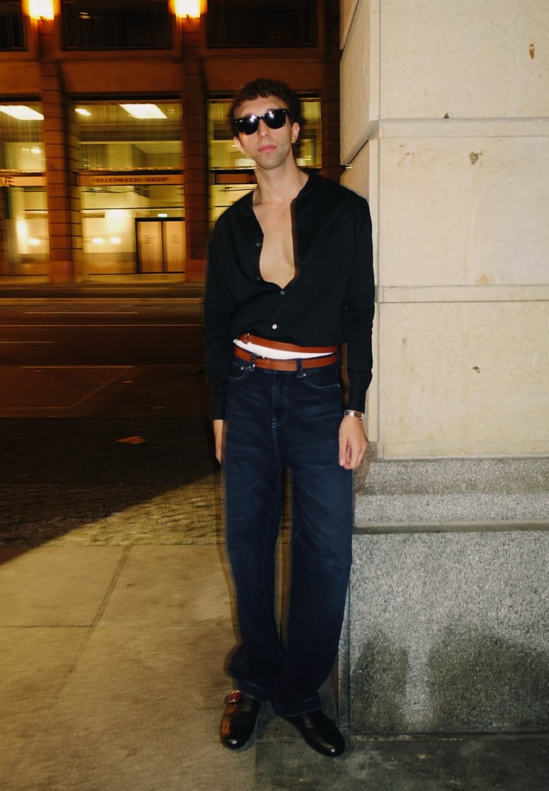 Camicia nera a bottoni, sbottonata sul petto, abbinata a jeans larghi blu scuro e cintura marrone. Occhiali da sole scuri e scarpe nere completano il look.
