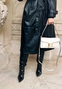 Trench coat in pelle nera con tasche laterali, abbinato a pantaloni in pelle nera attillati e stivali neri a punta. Tiene una piccola borsa bianca con hardware dorato.