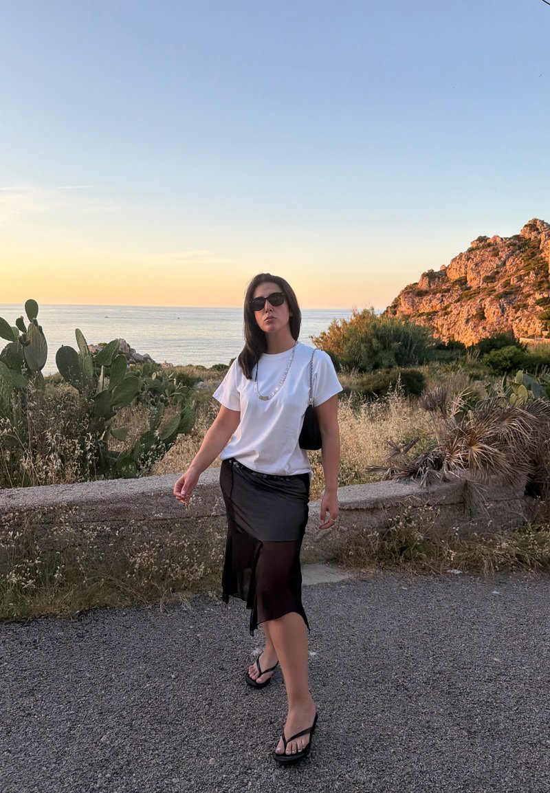 Femme portant des lunettes de soleil, une chemise blanche, une jupe noire et des sandales, marchant près d'une zone côtière avec des cactus et des collines rocheuses au coucher du soleil.