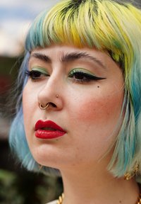 Κοντινό πλάνο ενός ατόμου με κοντά μαλλιά, κίτρινο-πράσινες ρίζες που ξεθωριάζουν σε μπλε, έντονο eyeliner, κόκκινα χείλη και έναν χρυσό δακτύλιο στη μύτη. Το δέρμα έχει φυσική υφή.