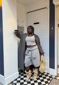 Sieviete ar pītām matu šķipsnām, valkā pelēku tanktopu, atbilstošus šortus un garu mēteli, bēšas zābaki, tur bēšu rokassomu, atspiedusies pret baltu sienu ārā.