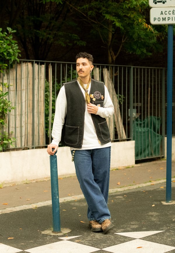Fekete mellény foltokkal, fehér hosszúujjú ing, kék bő szárú farmer és leopárdmintás cipő. Egy kék oszlop mellett áll.
