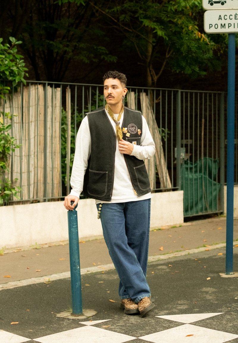 Colete preto com remendos, camisa de manga longa branca, jeans largos azuis e sapatos de padrão de leopardo. Em pé ao lado de um poste azul.
